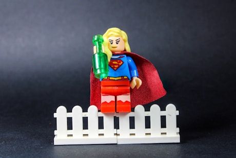 Supergirl lego figure holding a green bottle on fence.