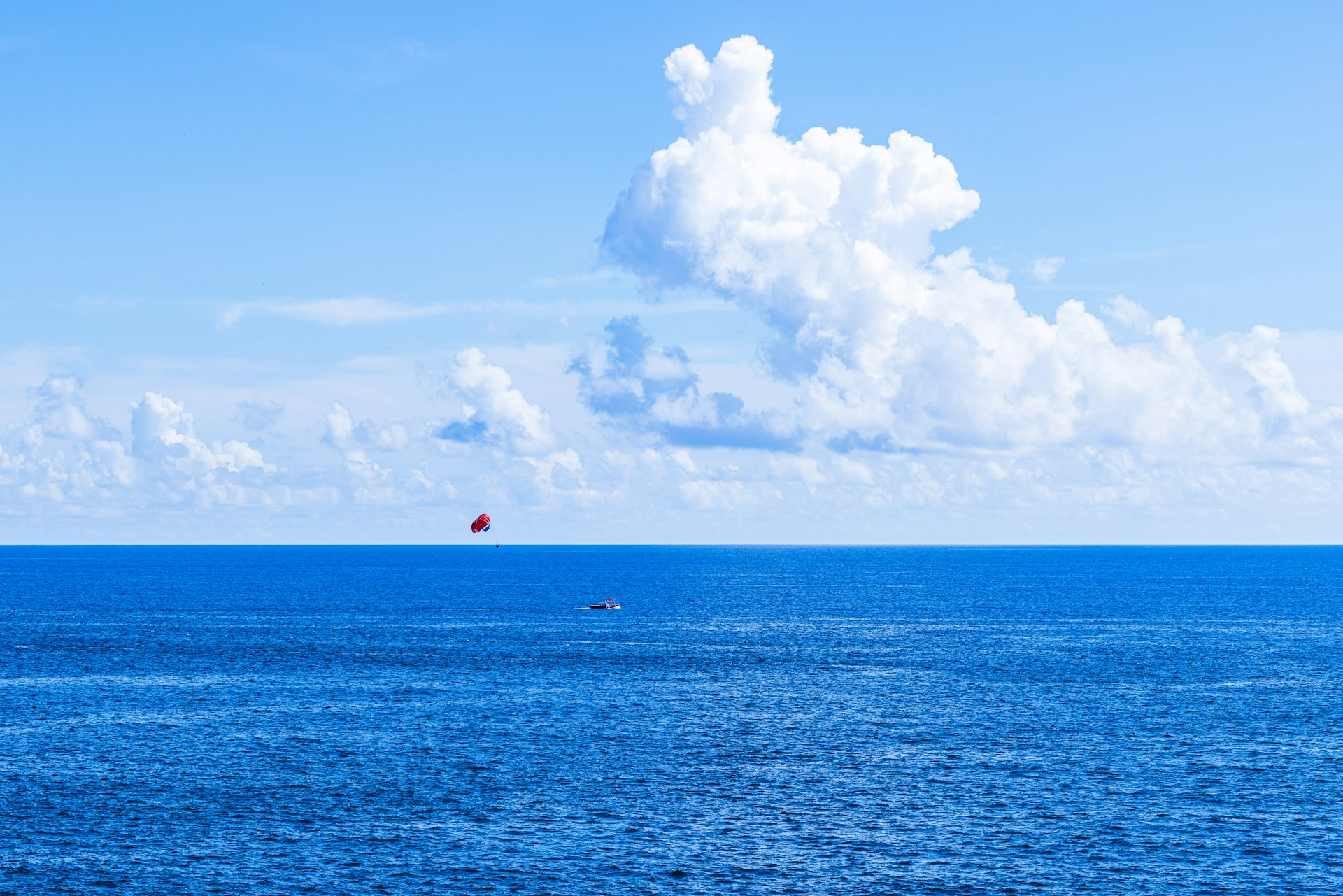 A red parasailer floats over the blue ocean under clouds.