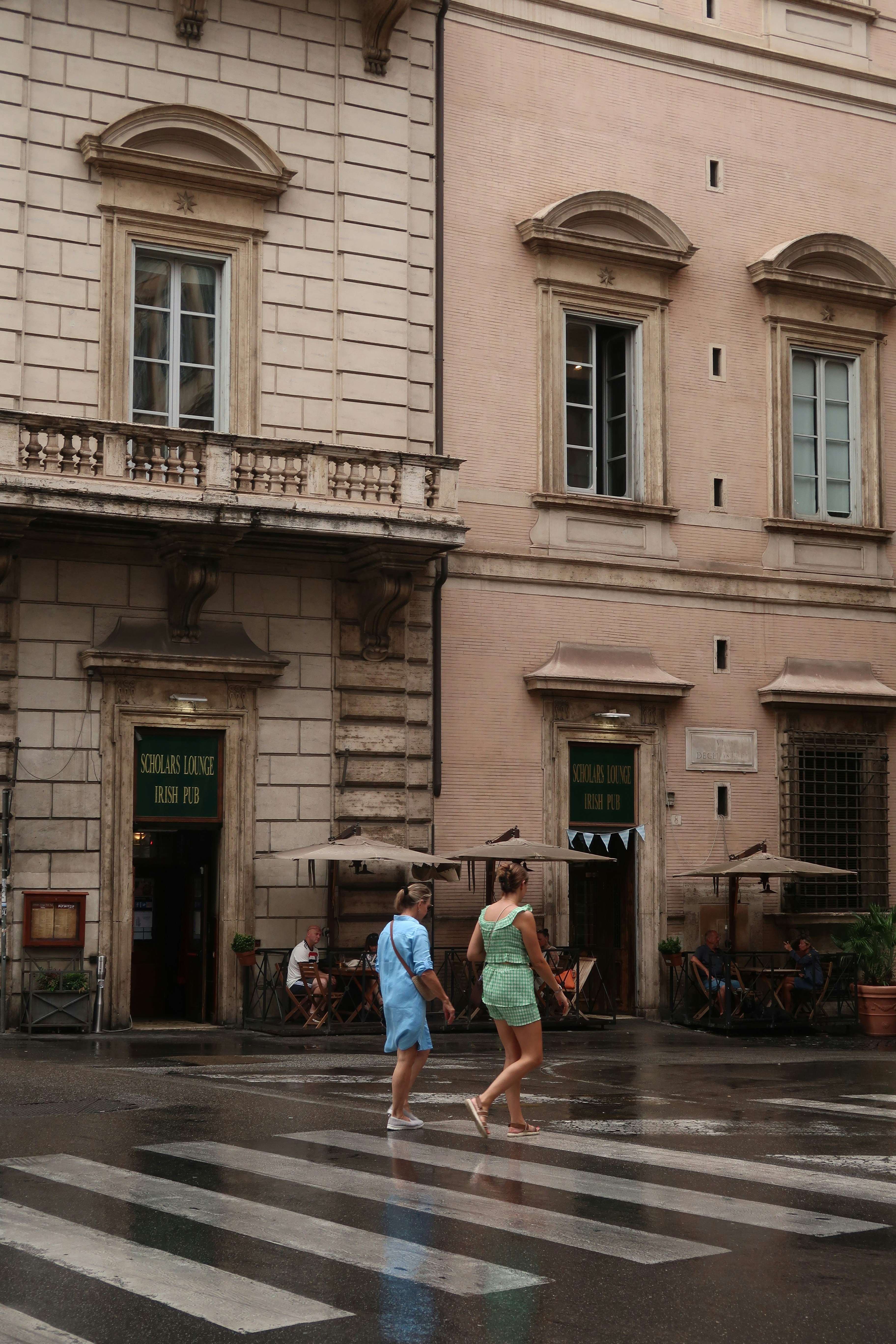 Two people cross a wet street in front of buildings.
