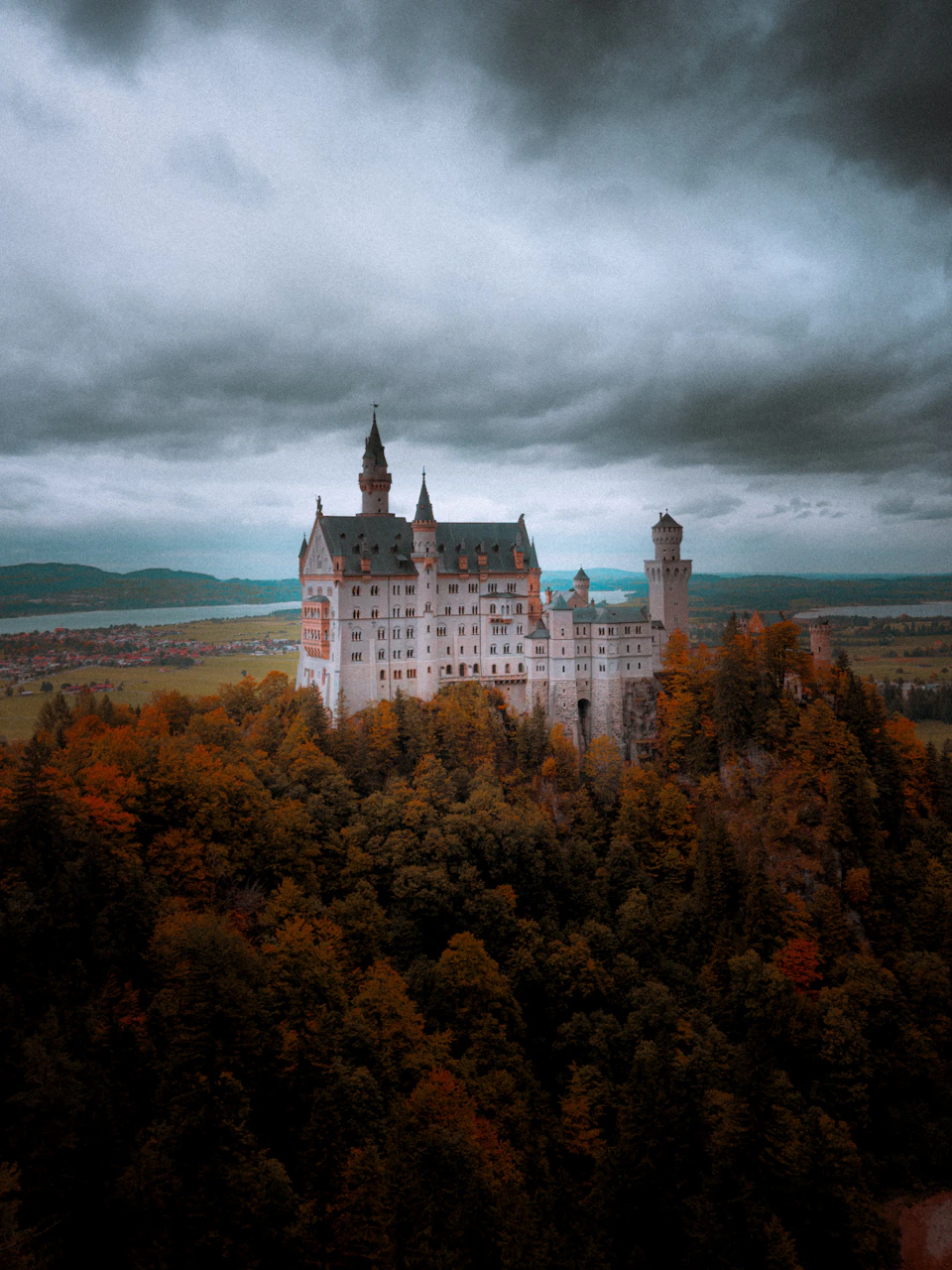 The image depicts a majestic castle perched on a hill, surrounded by lush greenery and trees. The castle features multiple towers and spires that reach towards the sky, creating an imposing presence against the backdrop of the cloudy day. The perspective of the photo is from below, looking up at the castle, which enhances its grandeur and majesty.