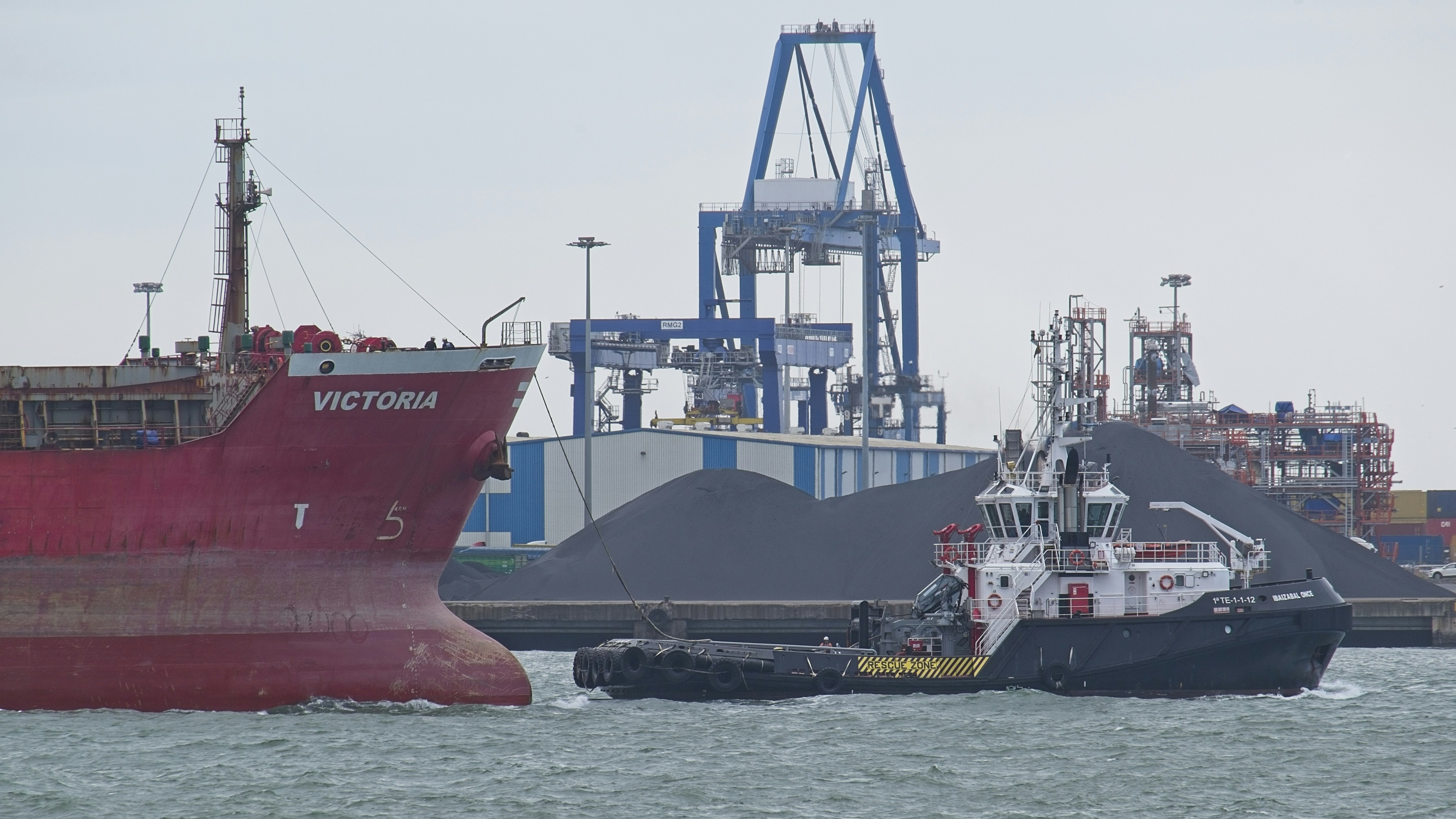Tugboat assists large red cargo ship in industrial harbor