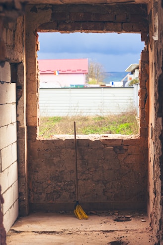 Broom leans against unfinished brick wall with window view.