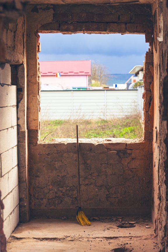 Broom leans against unfinished brick wall with window view.
