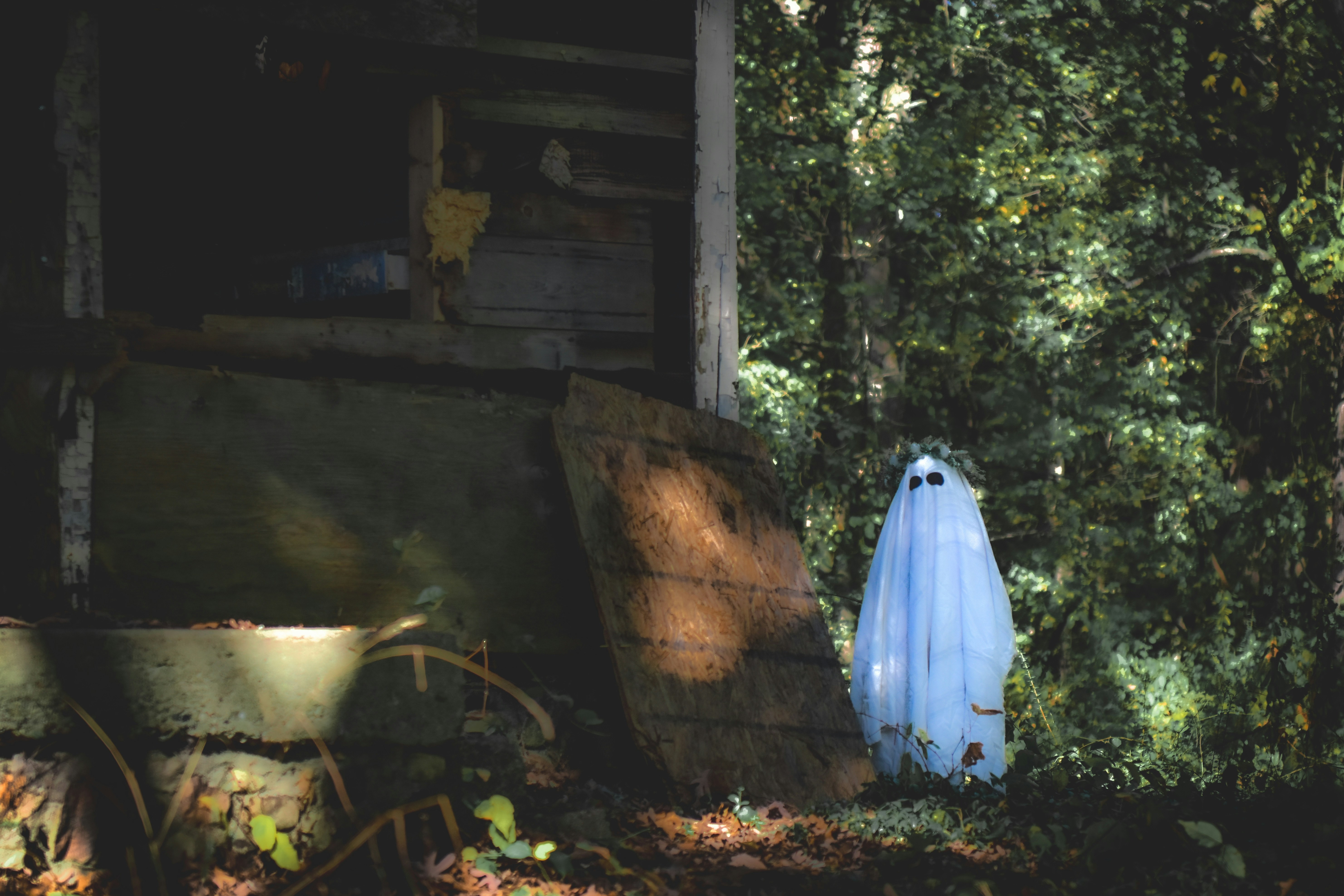 A ghost costume stands near a wooden structure in woods.
