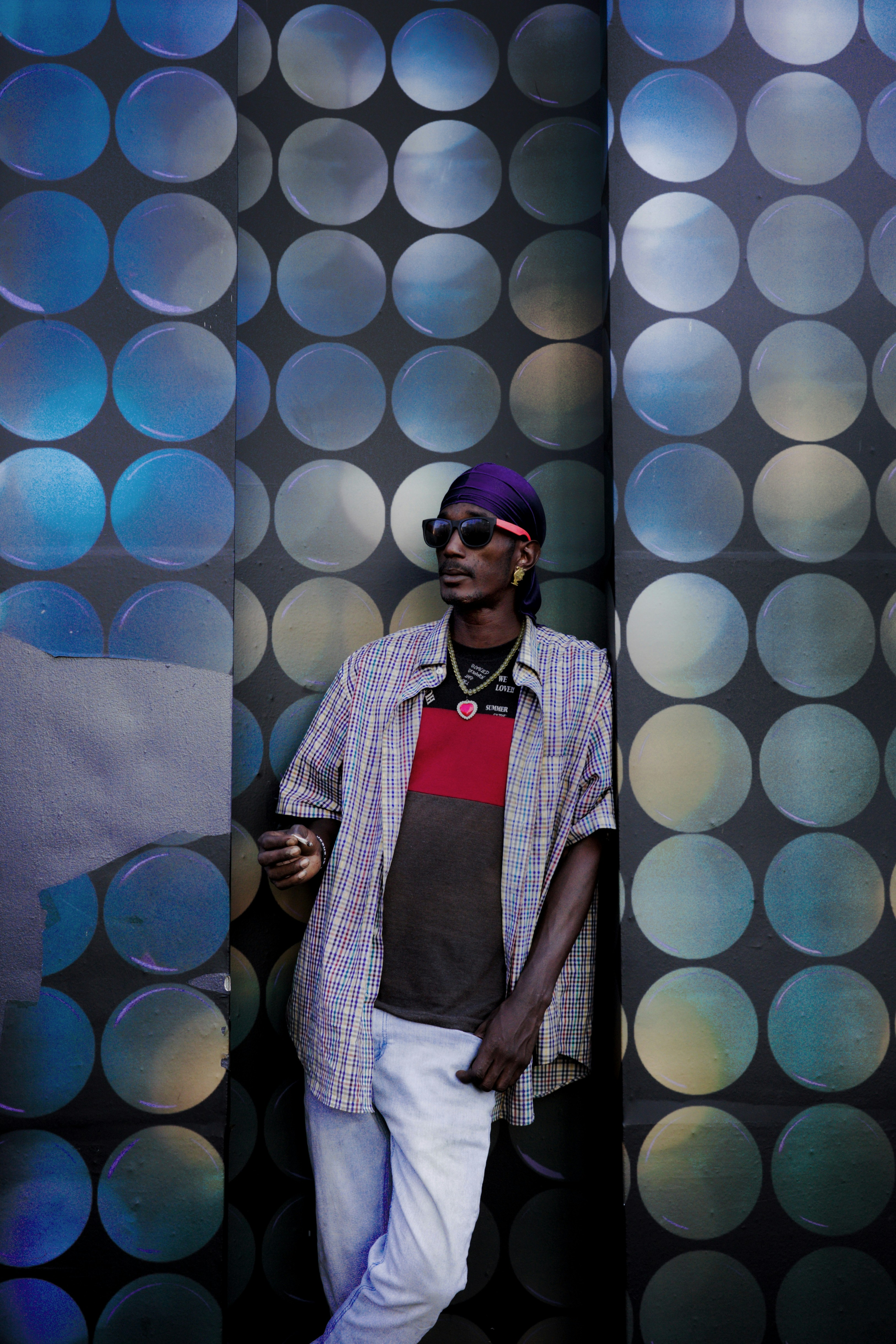 선글라스와 두건을 두른 남자가 무늬가 있는 벽을 배경으로 포즈를 취하고 있습니다.