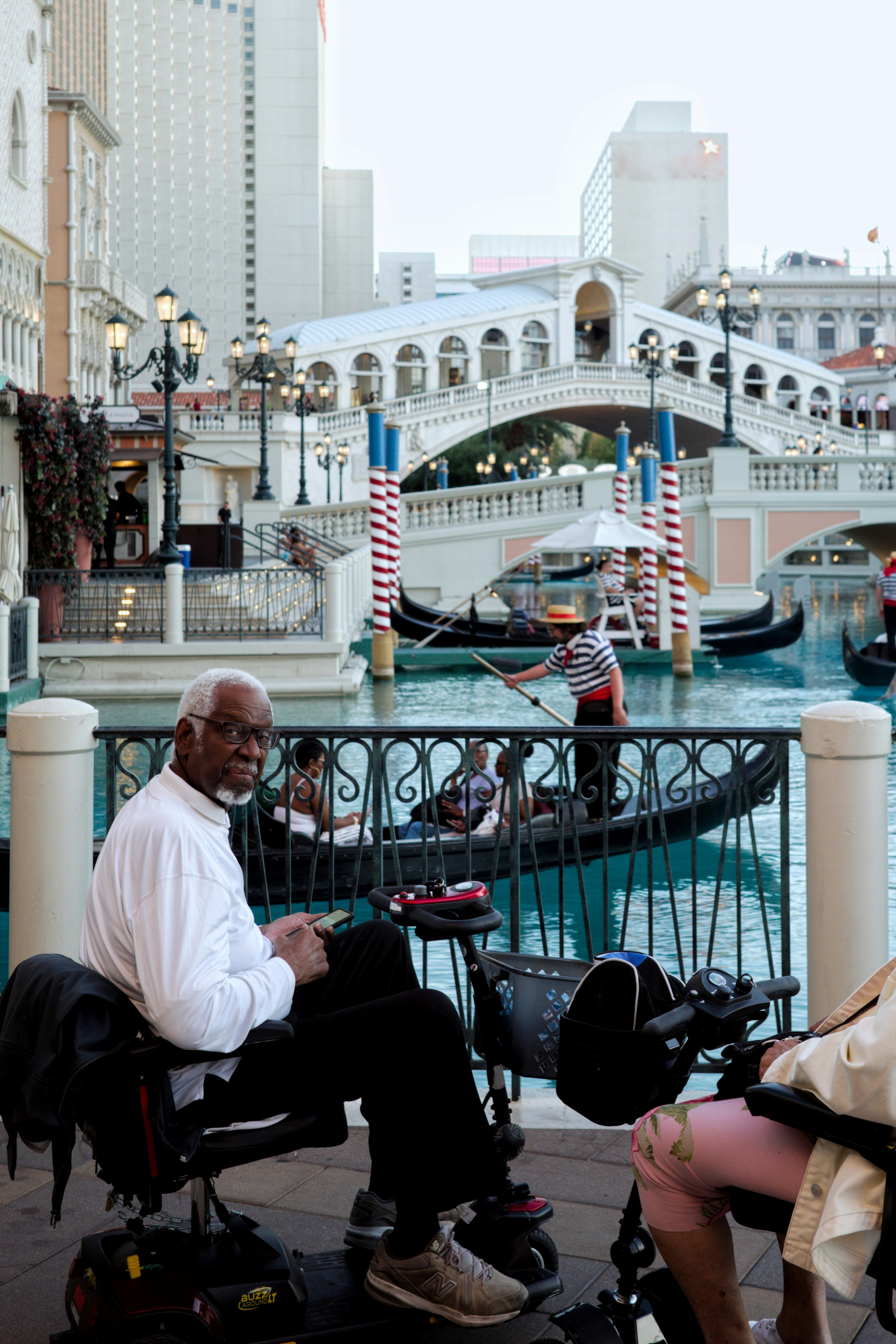 Man in wheelchair watches gondolas pass by canal.