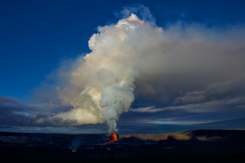 Tall plume of white steam and ash lit orange at its base rising from a dark crater floor at dusk