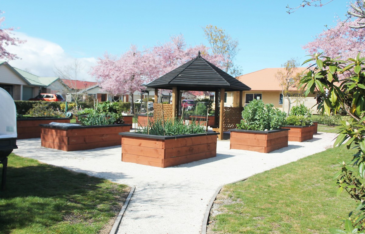 Raised garden beds with vegetables and flowers surround a gazebo in a community garden park.