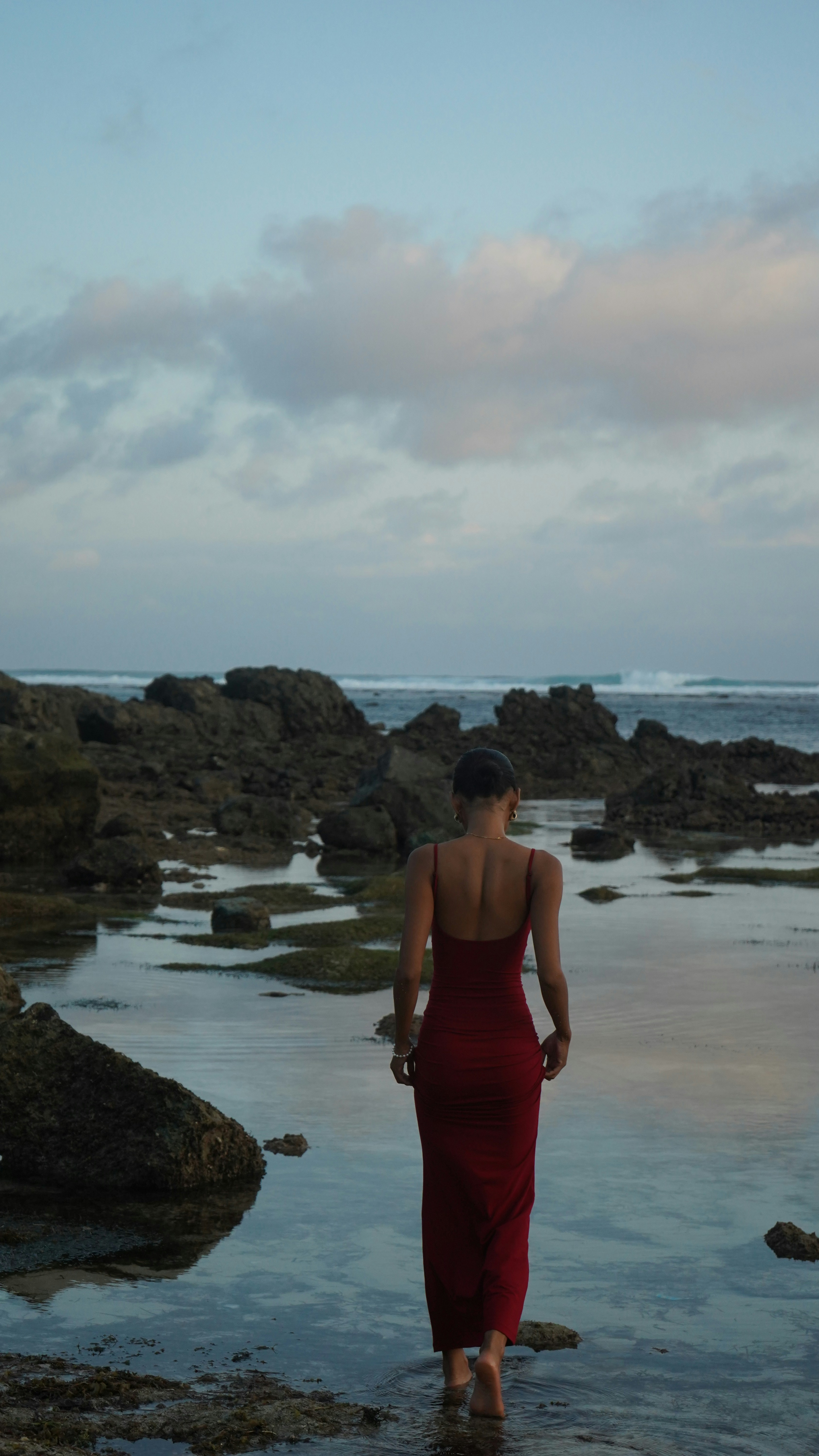 Woman in red dress walks on rocky shore at dusk