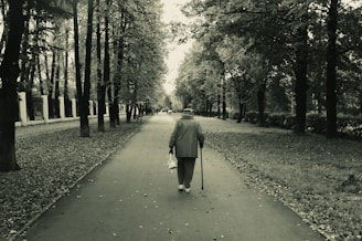 Elderly person walks down park path with cane.