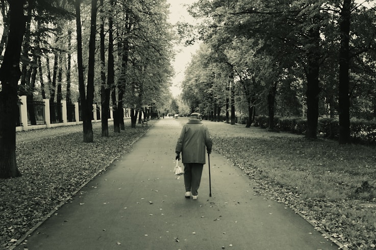 Elderly person walks down park path with cane.