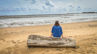 Woman sits on driftwood facing the ocean waves.
