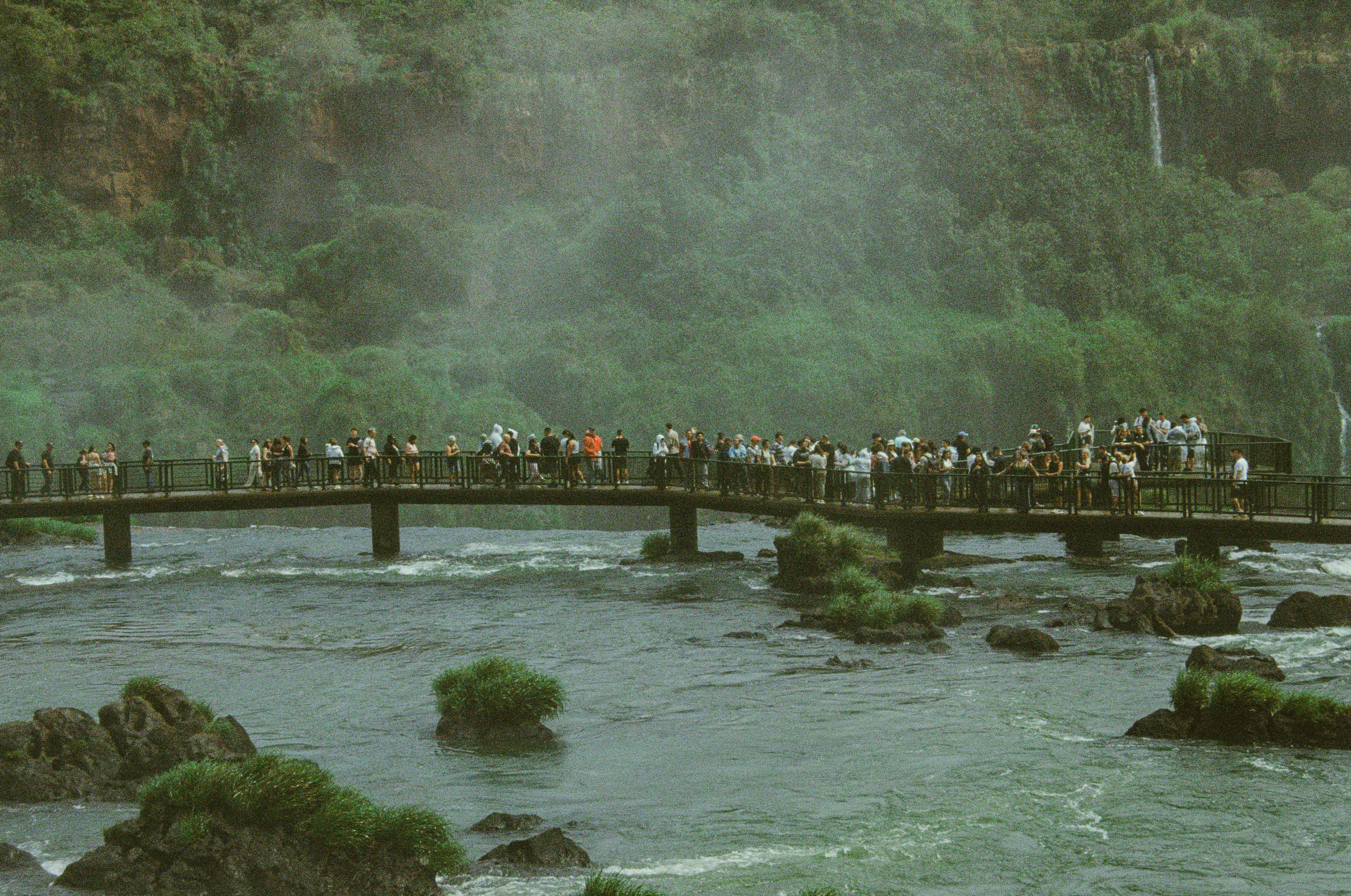 People walk on a bridge over a river near waterfalls.