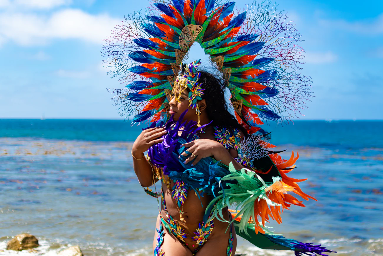 A performer in a vivid feathered carnival costume beside the ocean