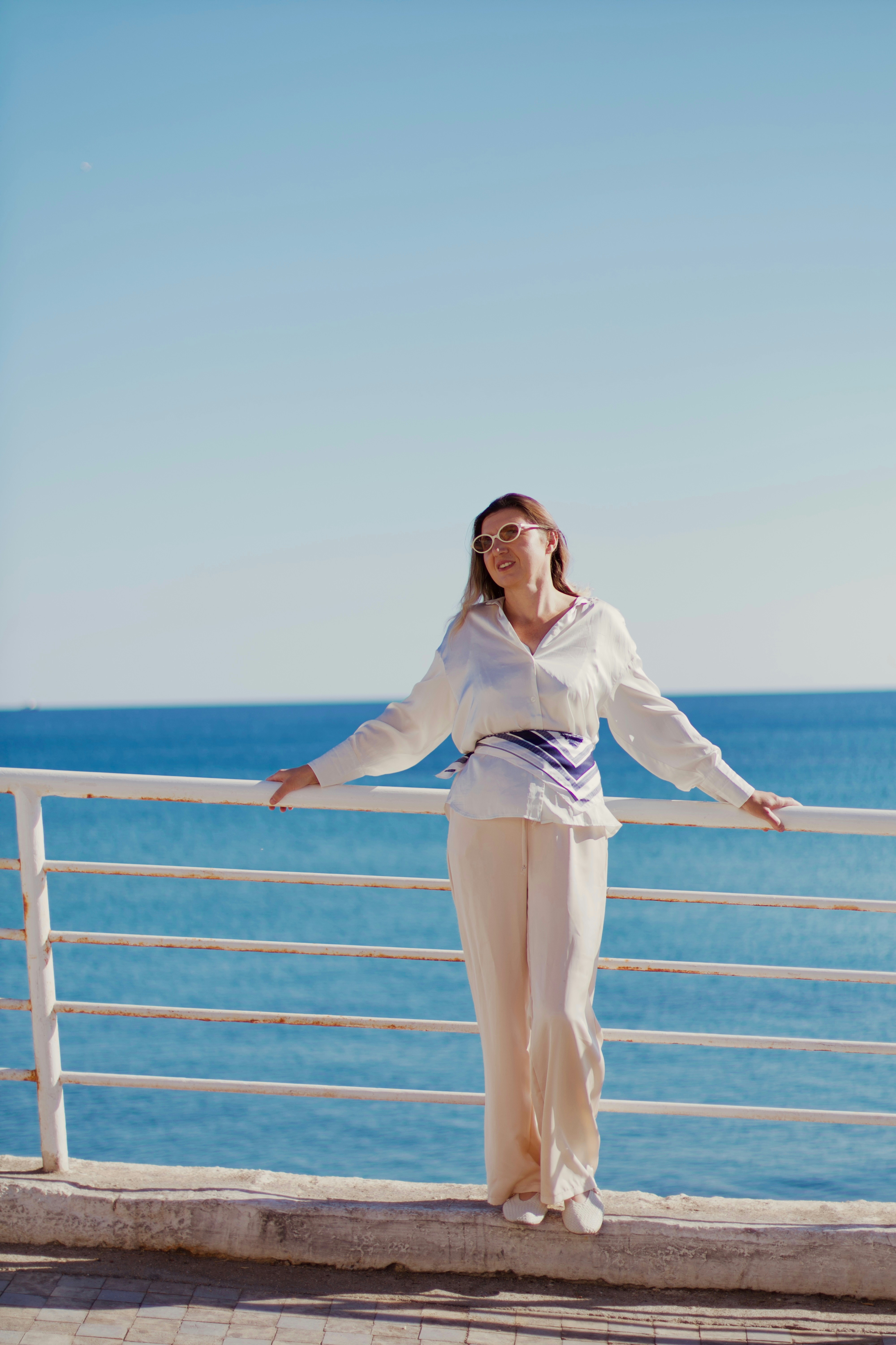 Woman in white outfit leans on railing by the sea.