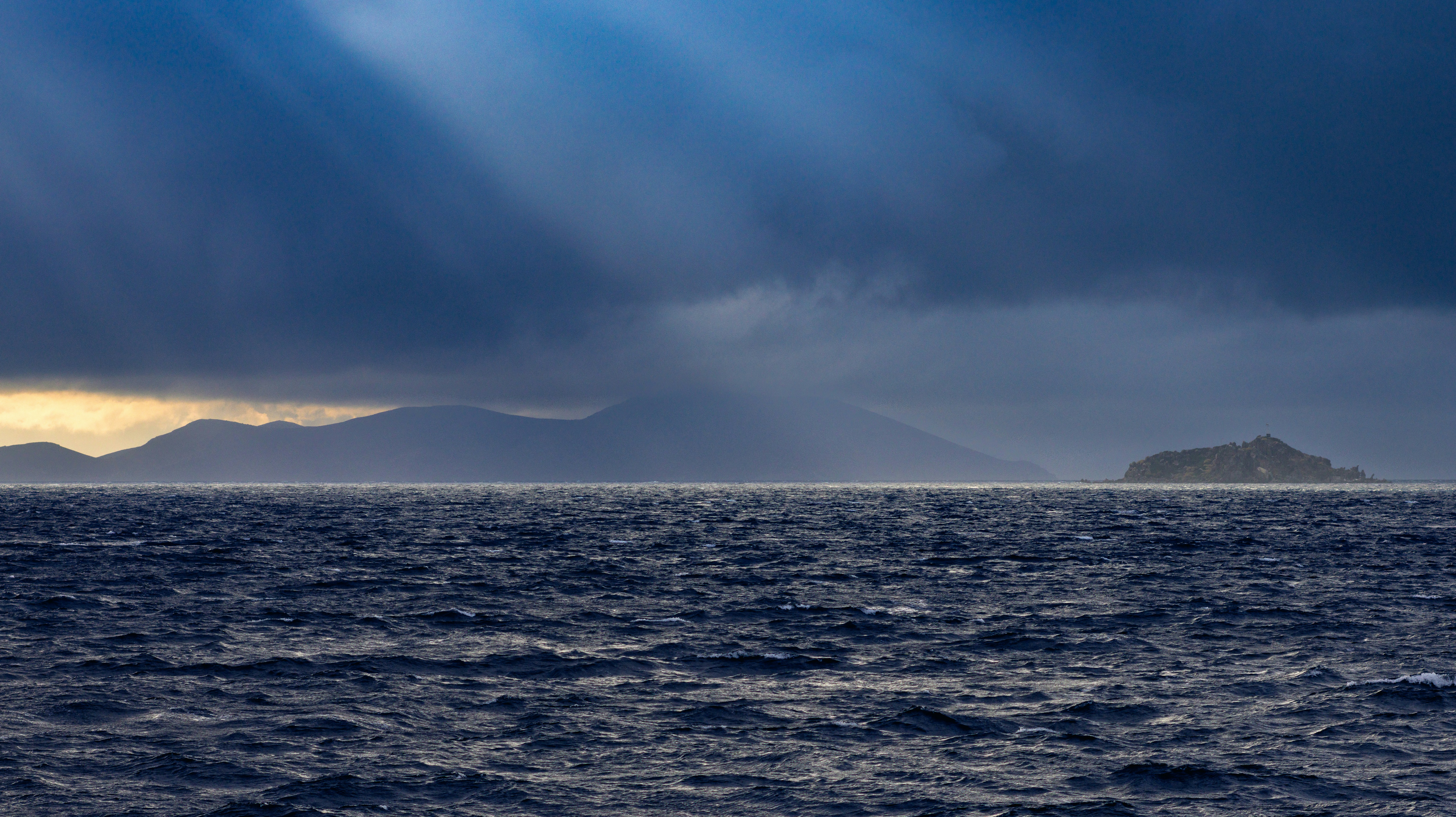 Dark stormy clouds over a choppy ocean with mountains.