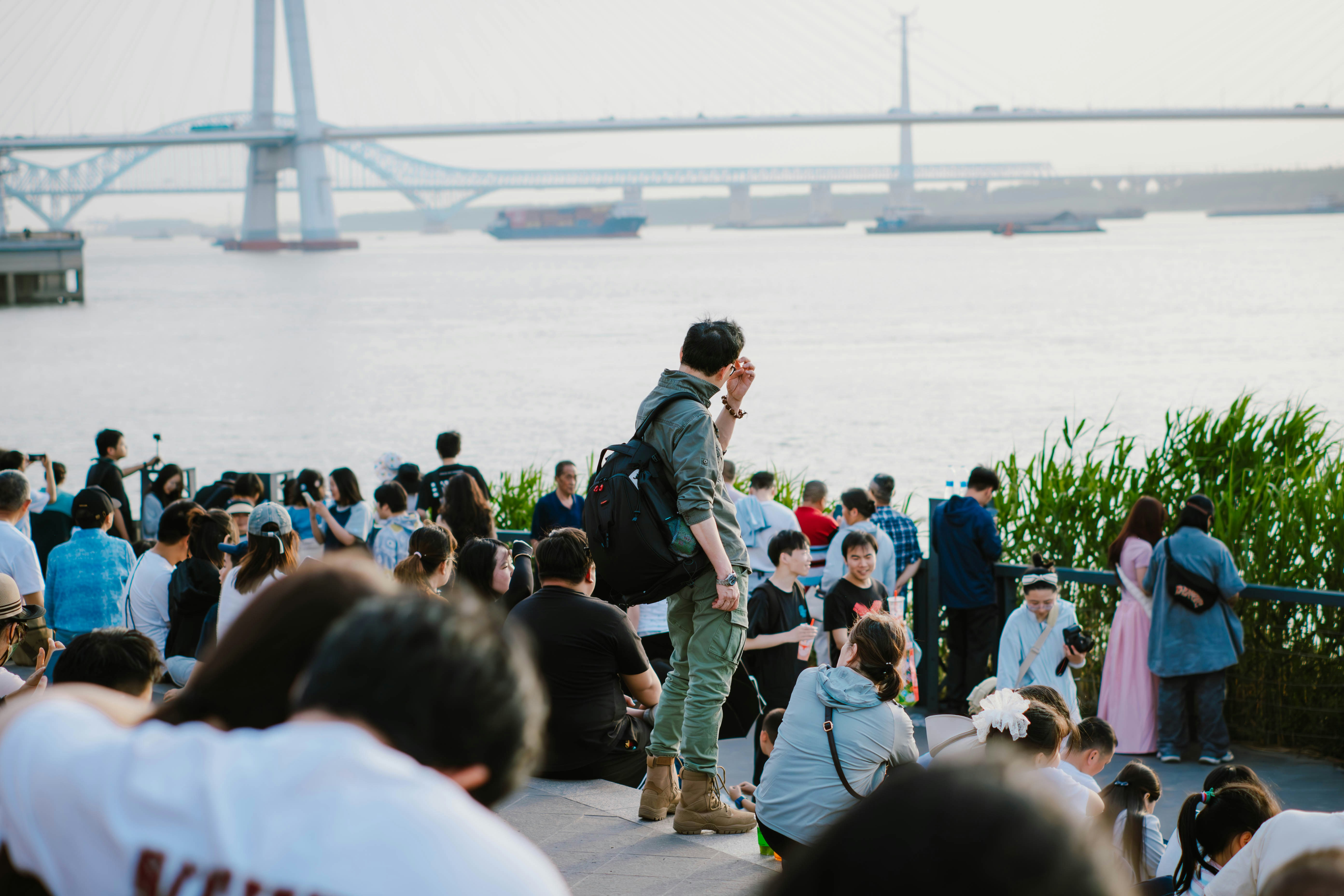 People gathered by a river with a bridge in background