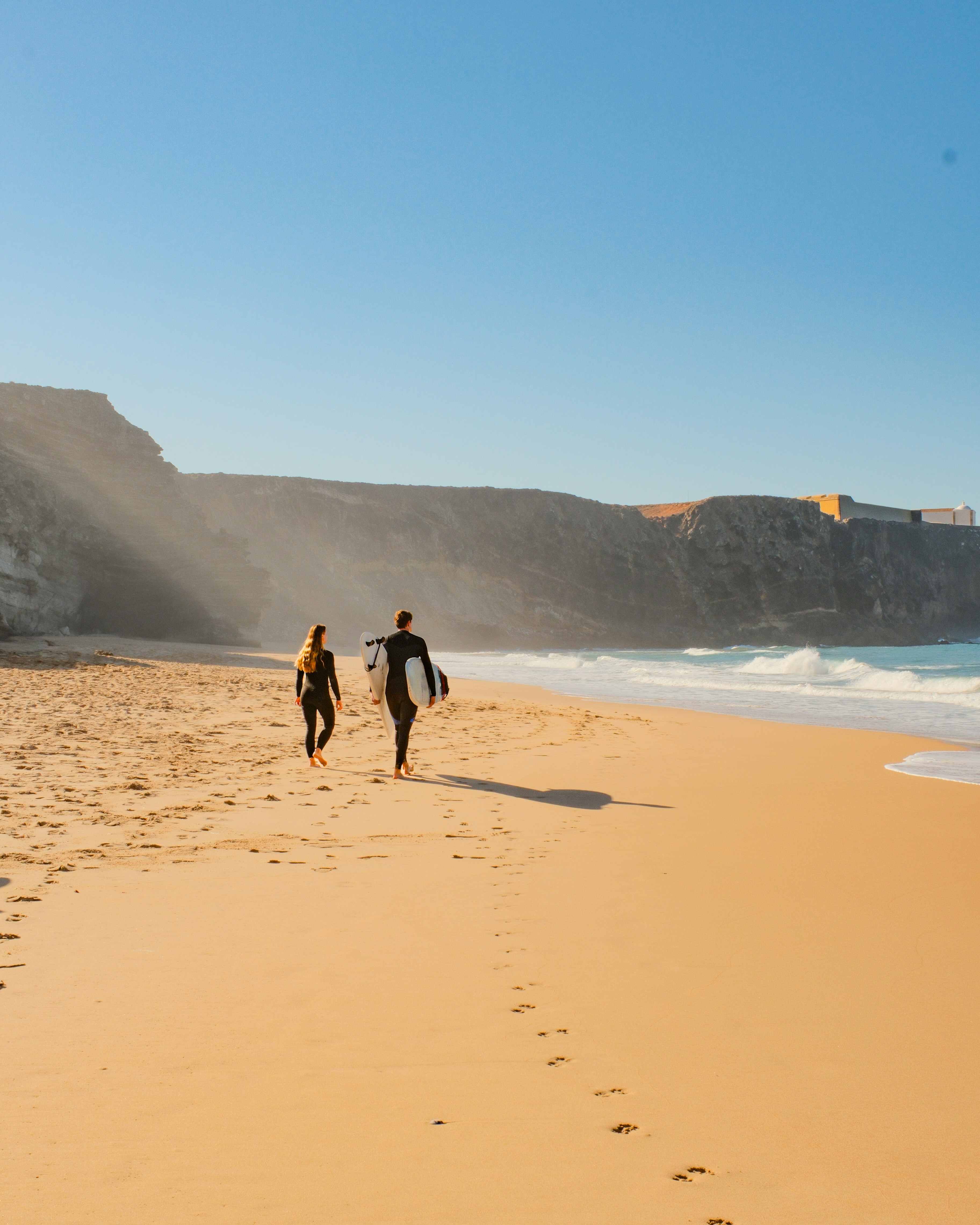 Two surfers walk along a sandy beach