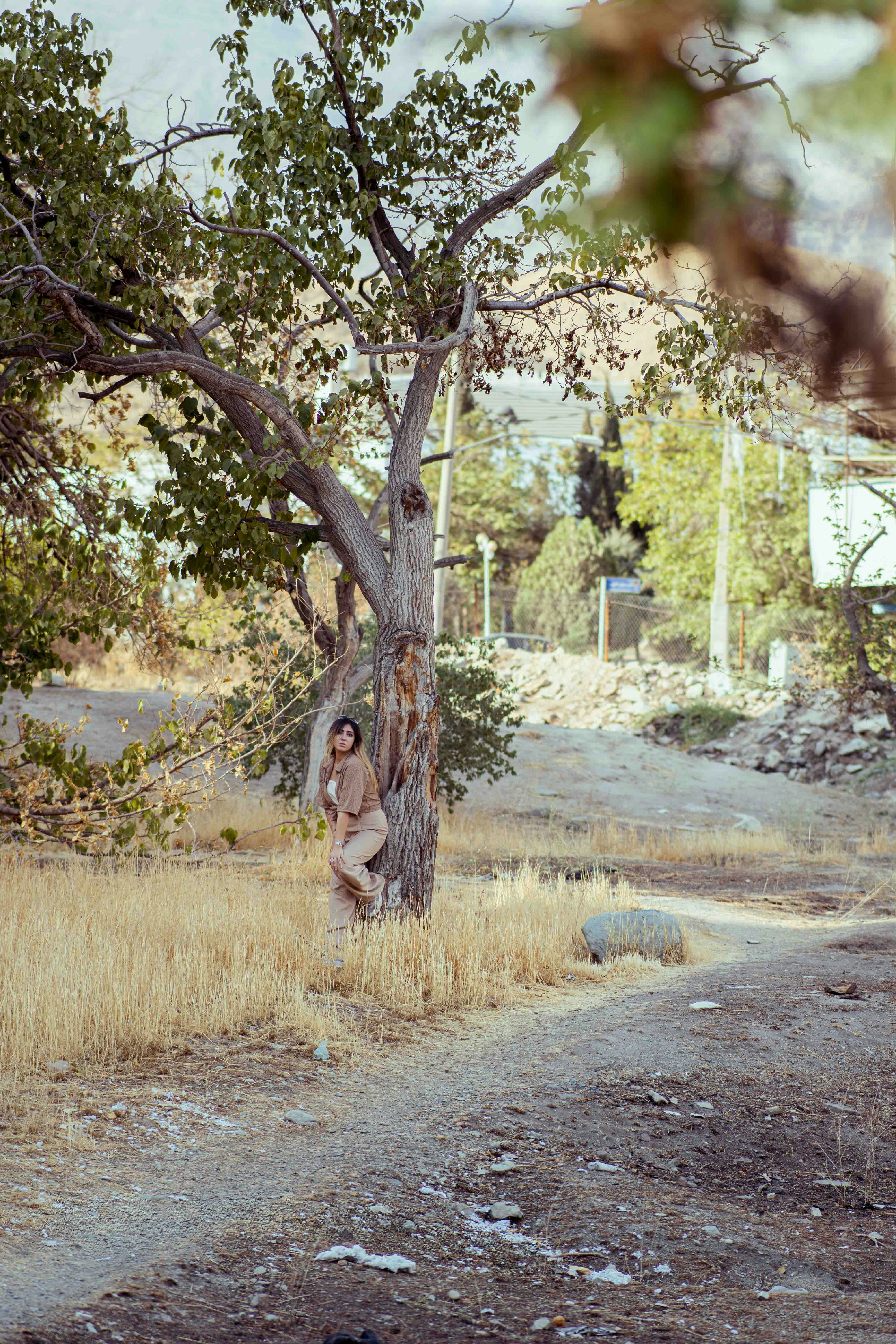 Woman leans against a tree in a dry, grassy field.