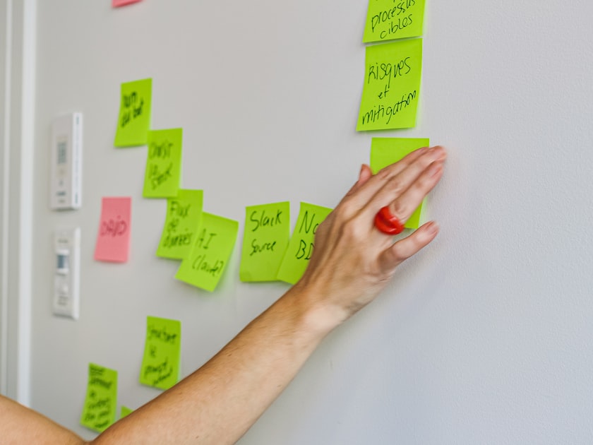 A hand placing a handwritten sticky note onto a wall covered in other notes, representing the manual work of coding customer language into repeated themes