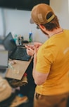 Man wearing yellow shirt and baseball cap using laptop.