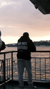 Man in "do not disturb" hoodie by water at sunset.