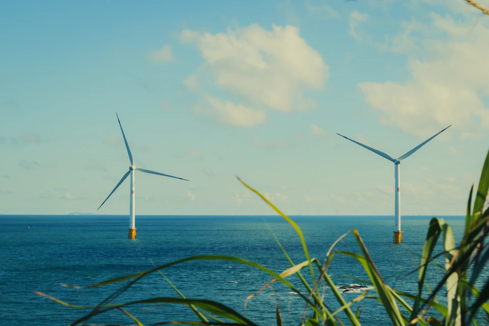 Two white wind turbines stand on rocky outcropping overlooking ocean with lush green grass below