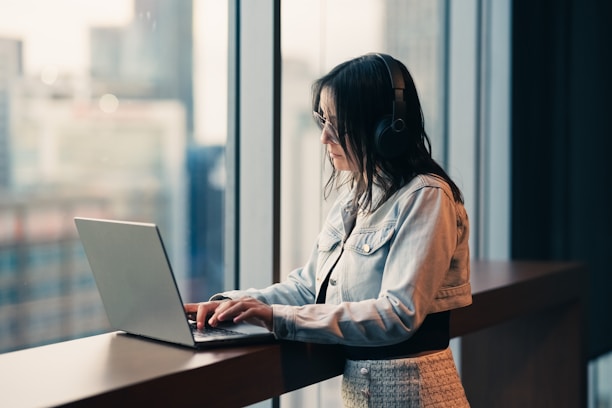 Woman wearing headphones works on a laptop by window.