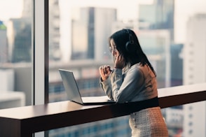 Woman wearing headphones works on laptop by window.