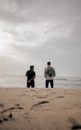 Two men stand on a sandy beach facing the ocean.