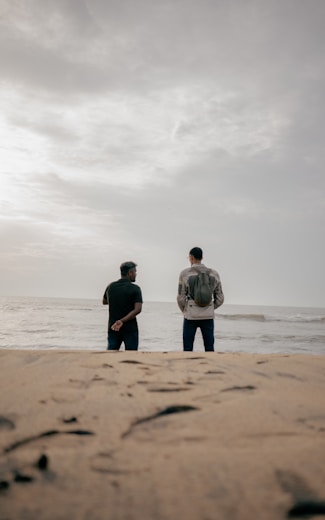 Two men stand on a sandy beach facing the ocean.