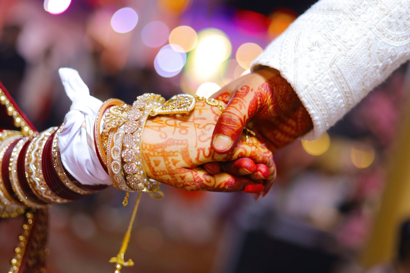 Close-up of a bride's henna-decorated hand held by the groom during a wedding ceremony with blurred lights behind
