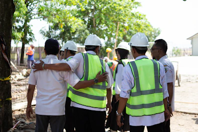 Construction workers in hard hats and vests huddle together.