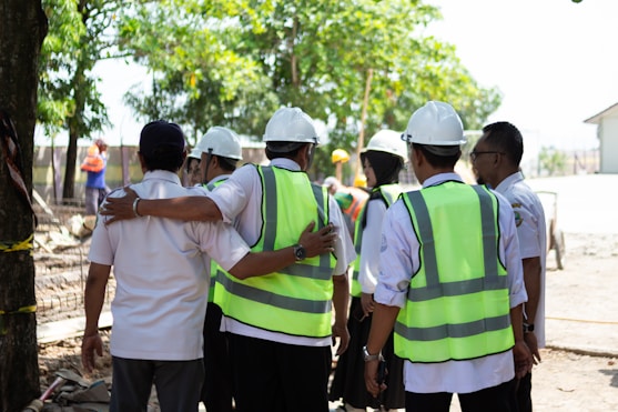 Construction workers in hard hats and vests huddle together.