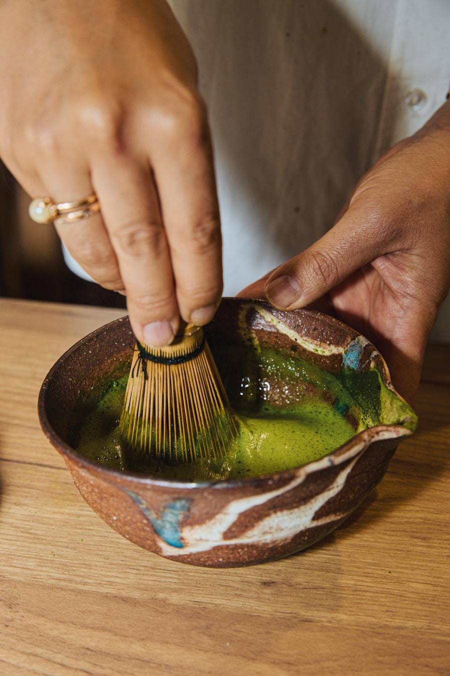 Hands whisking vivid green matcha with a bamboo whisk in a rustic ceramic bowl on a wooden table.