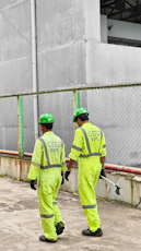 Two workers in neon safety suits walk past a fence.