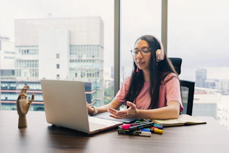 Woman wearing headphones talks at laptop by window.