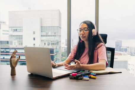 Woman wearing headphones talks at laptop by window.