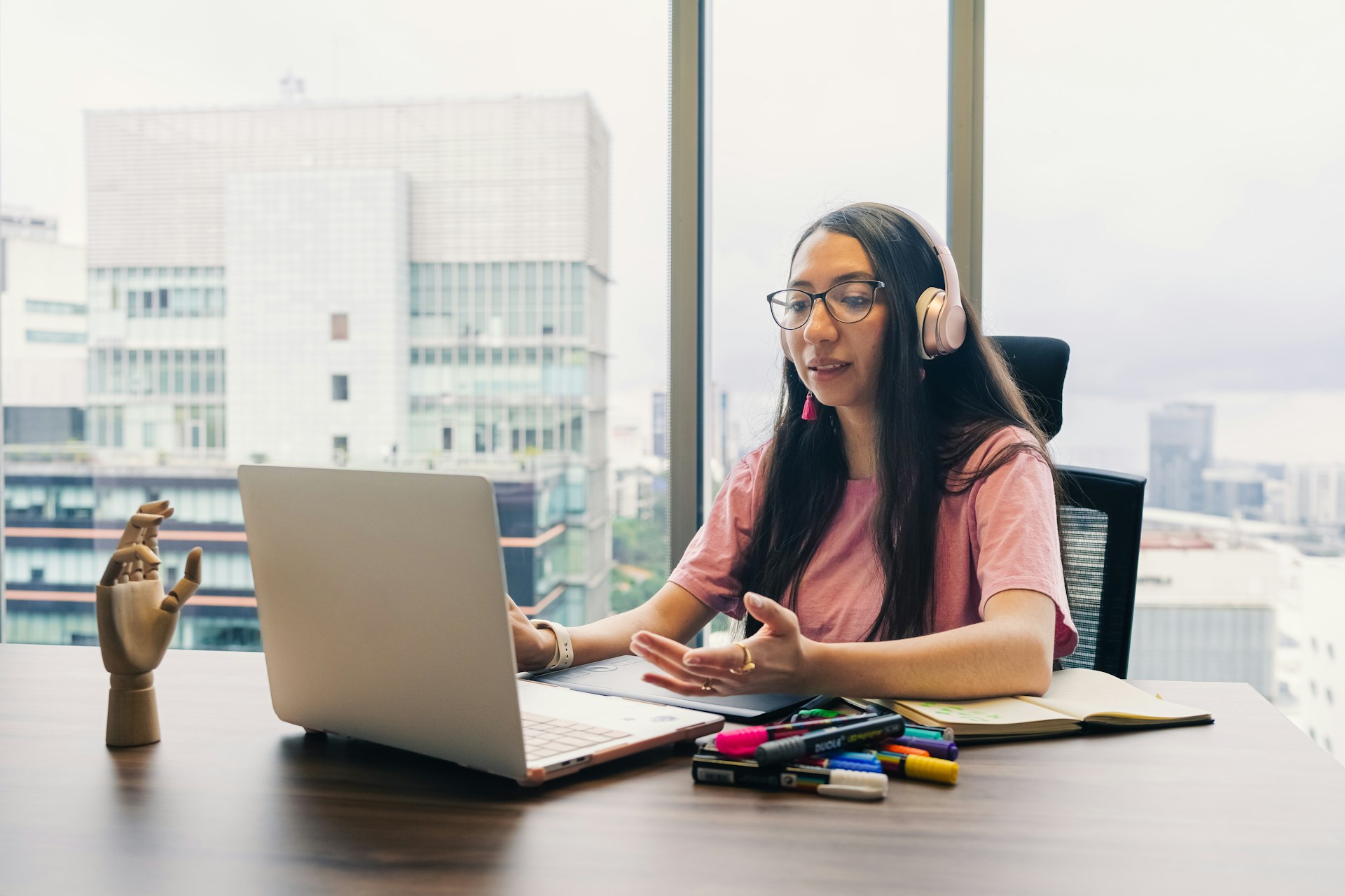 Woman wearing headphones talks at laptop by window.