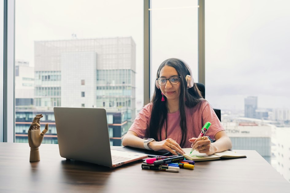 A woman in headphones and glasses sitting at a desk in front of a laptop during a video call, taking notes in a notebook with a highlighter