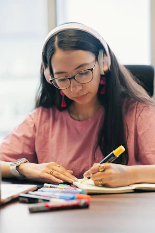 Young woman wearing headphones, glasses, and pink shirt writing.