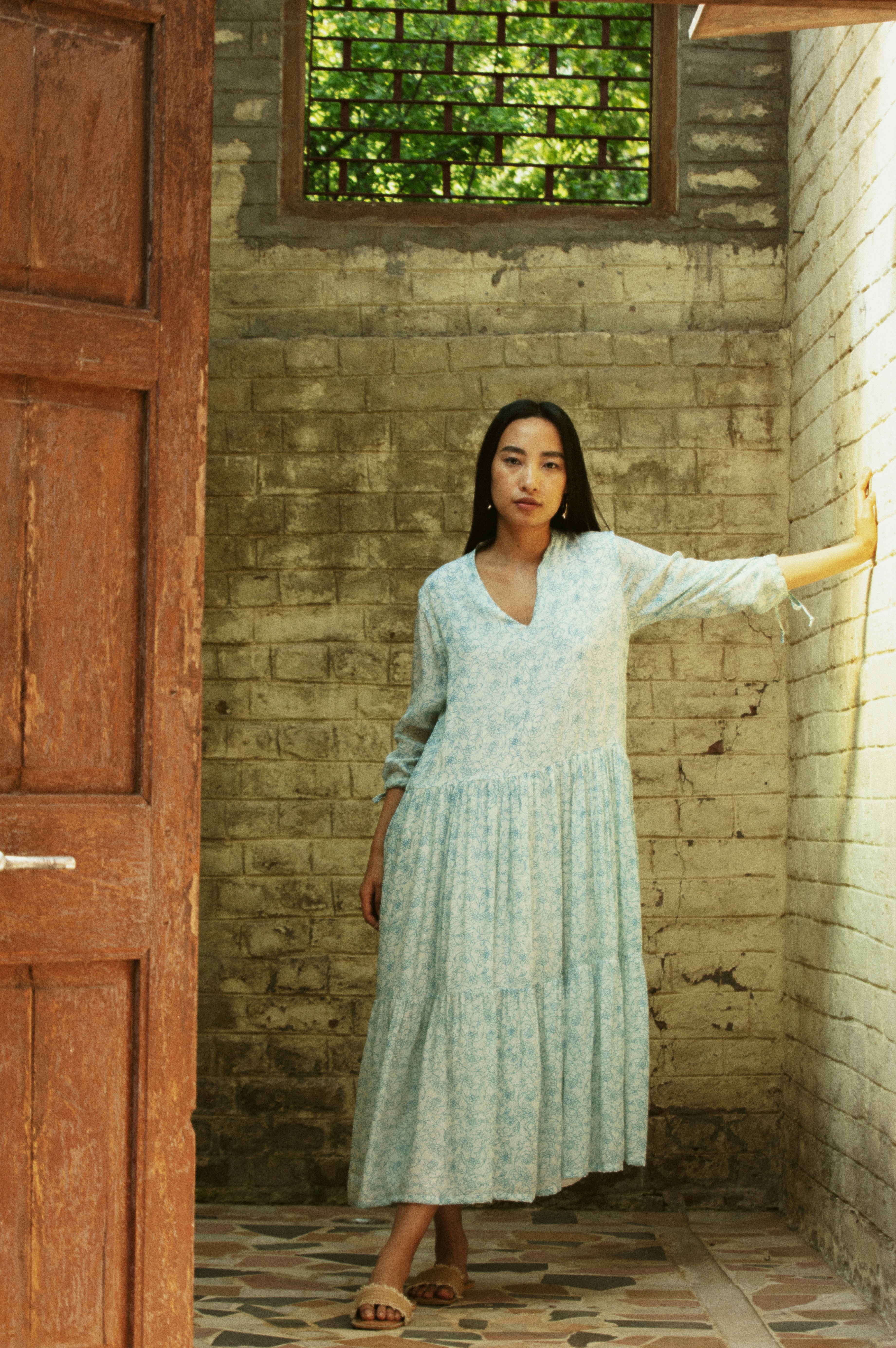 Young woman in a light blue dress leans against wall.