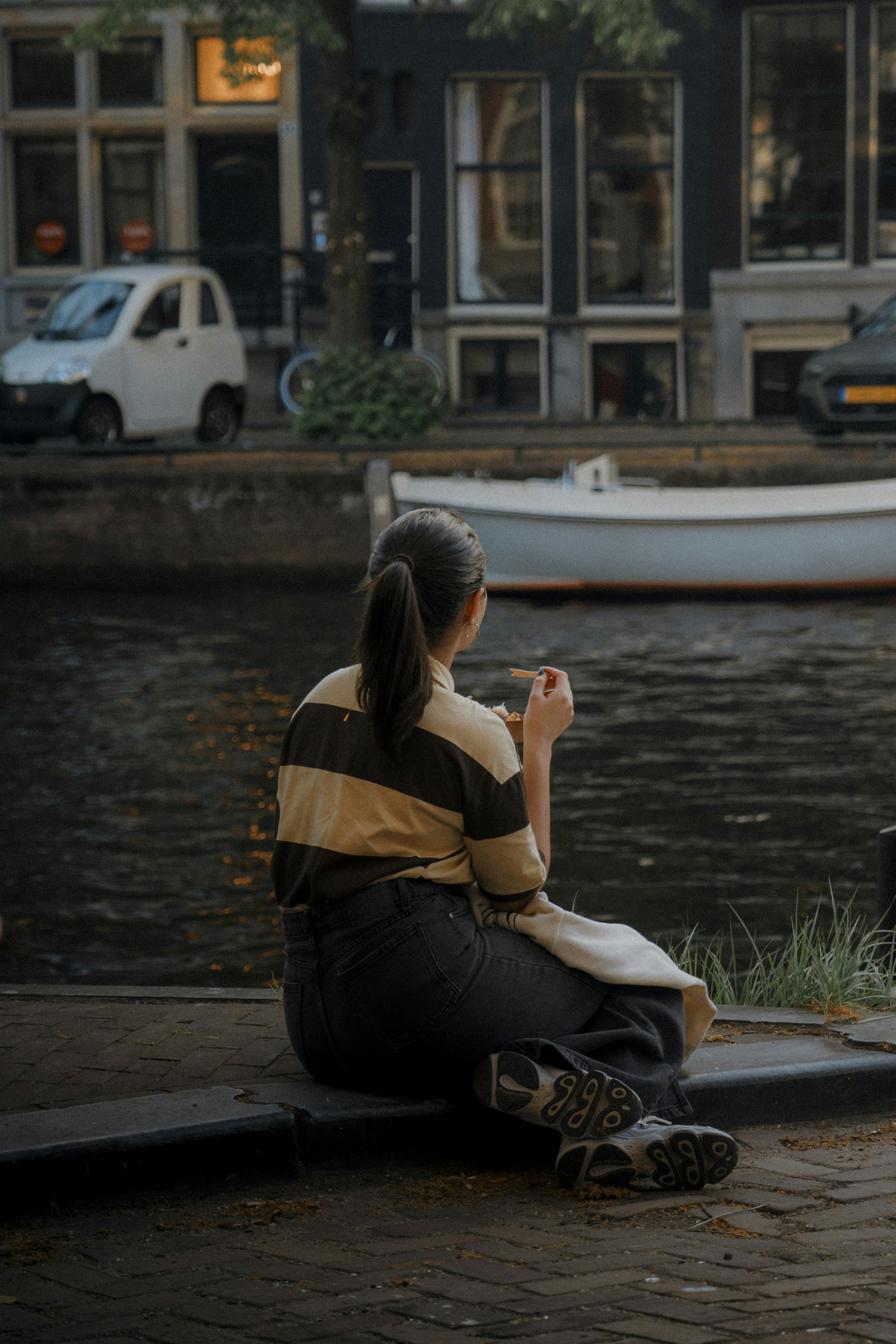 Woman sitting by canal with buildings and boat