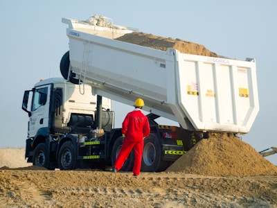 Worker in red supervises dump truck unloading sand