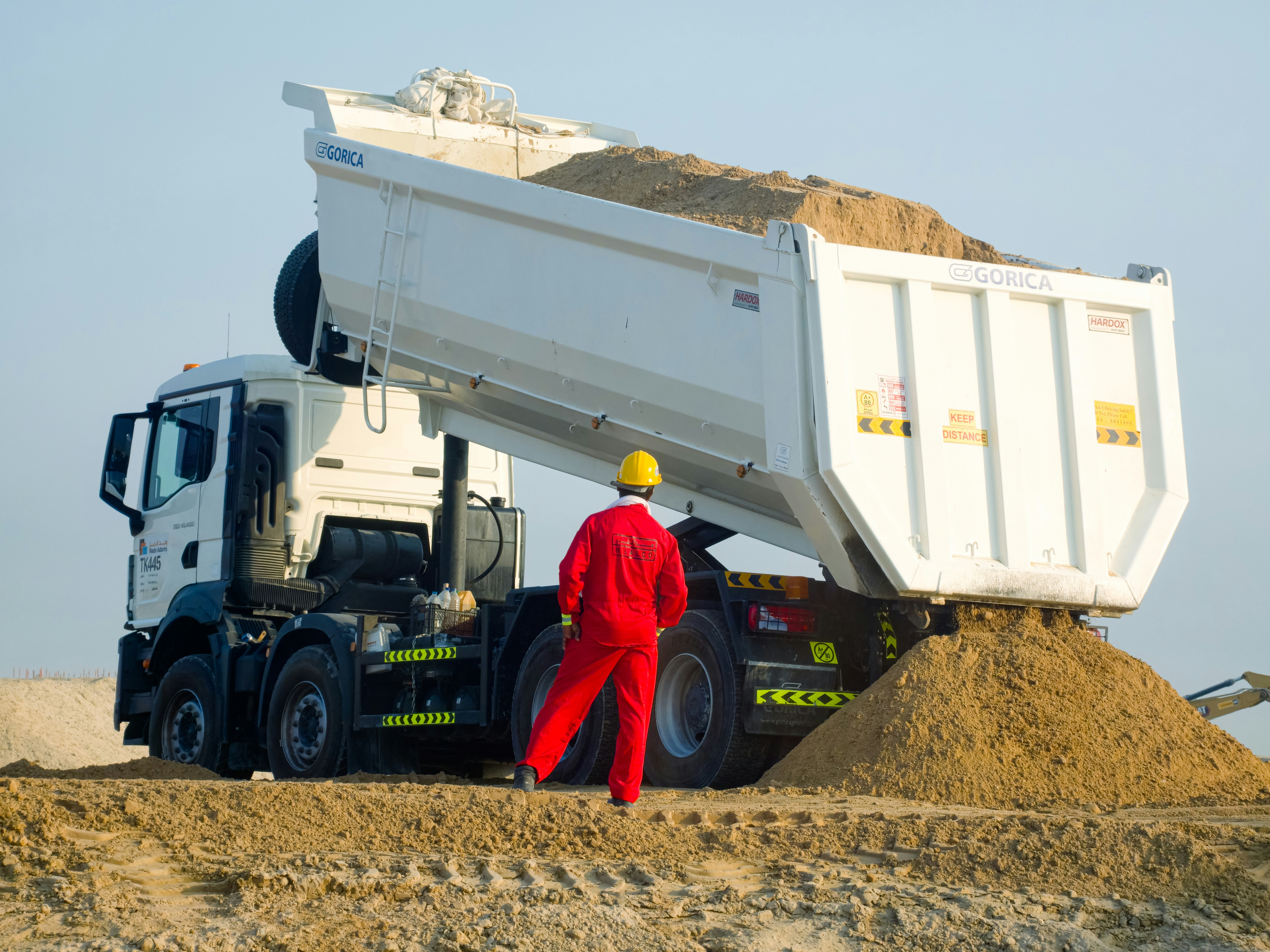 Worker in red supervises dump truck unloading sand