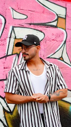Man in striped shirt and baseball cap against graffiti wall.
