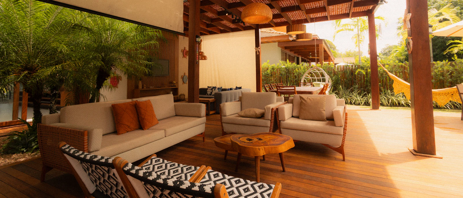 Covered tropical outdoor living area with timber decking, cream sofas and armchairs, patterned seating, a live-edge coffee table, woven lighting and lush landscaping.