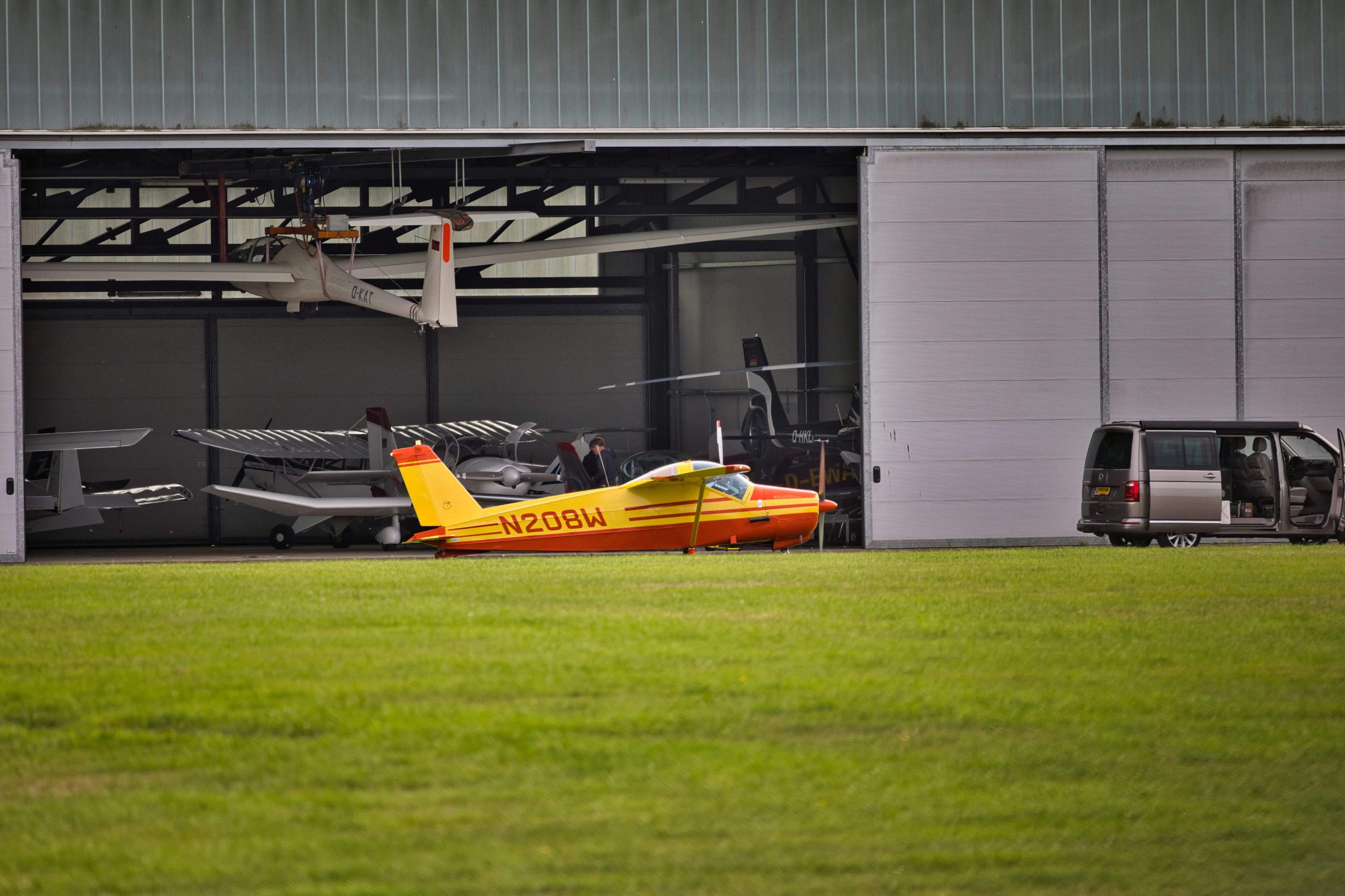 A small aircraft parked inside an open hangar beside a grass airfield.
