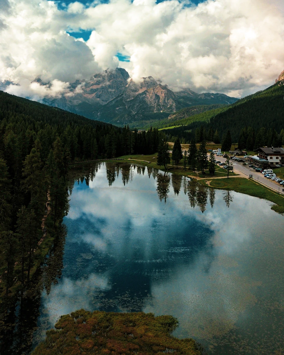 The image depicts a serene mountain lake nestled amidst lush green trees and rolling hills, with a small town visible on one side. The sky above is filled with fluffy white clouds, casting a soft light over the scene. The perspective of the photo is from an aerial view, providing a comprehensive look at the landscape below.