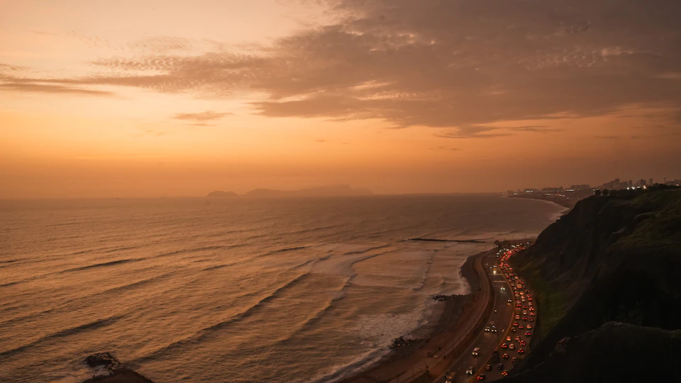 The image depicts a coastal scene at sunset, with a deep orange sky and light brown water reflecting the warm hues. The perspective is from a high vantage point, overlooking a highway that runs parallel to the shoreline. The highway is filled with cars, adding a sense of activity to the otherwise serene landscape. In the distance, several buildings can be seen on the cliffside, blending seamlessly into the natural surroundings.